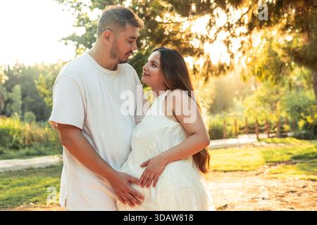 Un couple multinational profite d'un moment tendre dans un parc ensoleillé pendant la grossesse. Le cadre extérieur serein reflète l'amour et l'anticipation comme ils émettent Banque D'Images