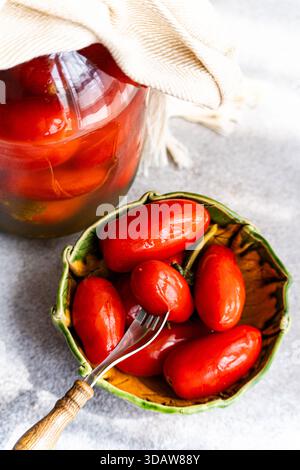 Un gros plan de tomates fermentées maison stockées dans un bocal en verre avec une couverture en tissu, à côté d'un bol rempli de tomates fermentées rouges vives sur un R. Banque D'Images