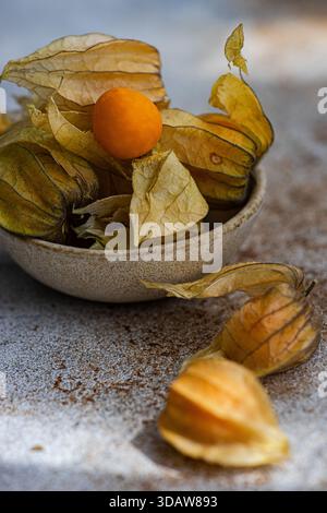 Un gros plan de physalis mûrs, également connu sous le nom de baie dorée ou groseille à maquereau du Cap, niché dans un bol rustique en céramique avec des feuilles séchées, mettant en valeur son éclat Banque D'Images