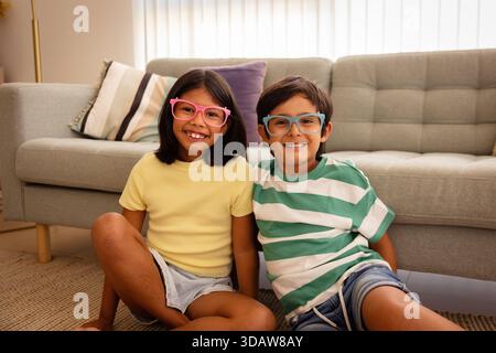 Deux enfants joyeux assis sur un sol confortable du salon, portant des lunettes lumineuses et colorées et des vêtements d'été décontractés. Ils sourient contre un COM Banque D'Images