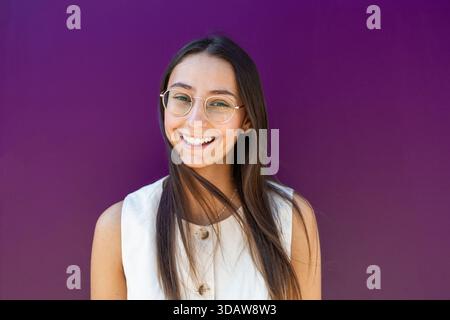 Une femme d'affaires joyeuse portant des lunettes sourit en toute confiance tout en se tenant devant un fond violet vibrant. Ses cheveux longs et sa tenue élégante R Banque D'Images