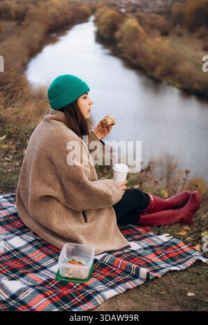 Une femme portant un bonnet vert et un manteau brun profite d'un pique-nique d'automne paisible au bord d'une rivière. Assise sur une couverture à carreaux, elle tient une tasse et une pâtisserie, sur Banque D'Images