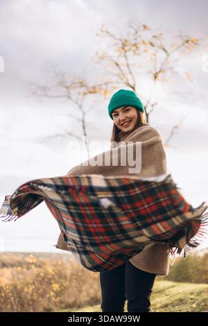 Une femme dans un bonnet et enveloppée chaleureusement dans une couverture marche joyeusement à travers un paysage d'automne, profitant de l'air frais et du feuillage coloré. Sa joie Banque D'Images