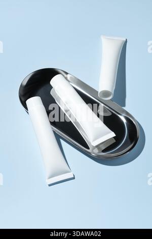 Trois tubes cosmétiques blancs de crème reposent élégamment sur un plateau métallique élégant, sur un fond bleu apaisant. Cette maquette minimaliste est parfaite Banque D'Images