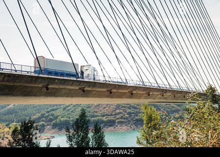 Un transport de marchandises se déplace sur un pont à haubans sous un ciel dégagé, avec pour toile de fond un paysage vibrant comprenant une rivière et des collines verdoyantes, Banque D'Images