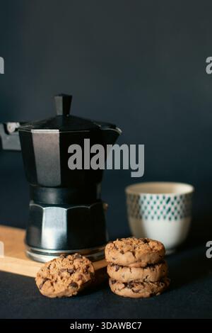 Un arrangement appétissant de biscuits faits maison à côté d'une cafetière vapeur et d'une tasse élégante sur un fond sombre, créant un mor confortable et accueillant Banque D'Images