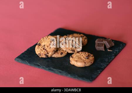 Un lot de délicieux biscuits aux pépites de chocolat maison repose séduisamment sur une assiette d'ardoise noire sur un fond chaud et rouge, accompagné d'un petit choc Banque D'Images