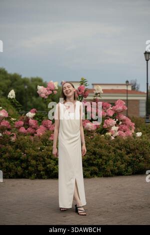 Une femme portant une longue robe blanche se tient joyeusement dans un jardin entouré de fleurs vives roses et blanches en fleurs, créant un calme et joyeux Banque D'Images