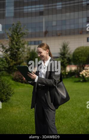 Femme d'affaires en tenue professionnelle tient une tablette tout en souriant à l'extérieur dans un cadre urbain. Elle se tient sur une zone herbeuse, avec des bâtiments modernes en th Banque D'Images