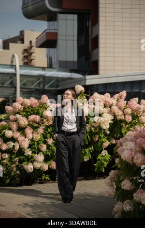 Femme d'affaires confiante dans un costume sur mesure se promène dans un jardin urbain, entouré de fleurs luxuriantes, dans un contexte d'architecture urbaine moderne Banque D'Images