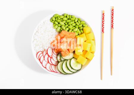 Vue de dessus d'un bol de poke vibrant avec saumon frais, riz, edamame, radis, concombre, et mangue sur fond blanc, accompagnée de baguettes. Banque D'Images