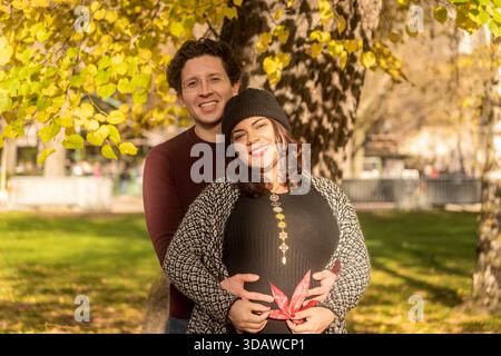 Un joyeux couple enceinte embrasse sous des feuilles d'automne dorées. La femme, portant un bonnet, sourit tout en tenant une feuille rouge sur son ventre comme l'homme l Banque D'Images