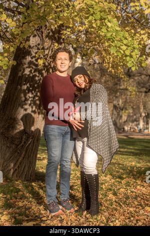 Une femme enceinte souriante se tient avec son partenaire dans un parc d'automne, embrassant sous un arbre avec des feuilles vibrantes, symbolisant l'amour et l'anticipation pour Banque D'Images