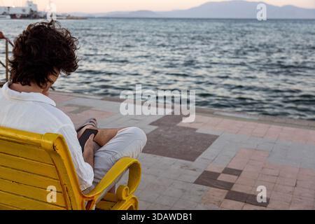 Un jeune homme est assis sur un banc jaune près de la plage, engrossé dans son smartphone. L'océan serein et un coucher de soleil coloré créent un outd tranquille et pittoresque Banque D'Images