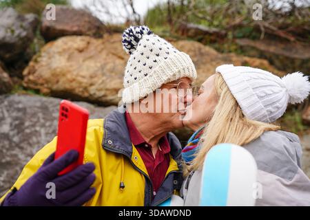 Un couple plus âgé partage un moment joyeux en plein air pendant la saison hivernale. Les deux sont chaudement habillés, capturant un selfie alors qu'ils embrassent le froid Banque D'Images