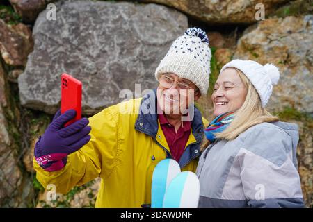 Un couple plus âgé profite d'une agréable journée d'hiver, chaudement vêtu de vestes et de bonnets. Ils capturent l'instant avec un selfie joyeux, entouré de bea Banque D'Images