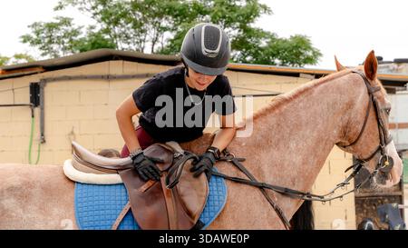 Une jeune femme portant un casque et des gants monte un cheval sellé dans un centre équestre. La scène capture un moment de préparation et de connexion Banque D'Images