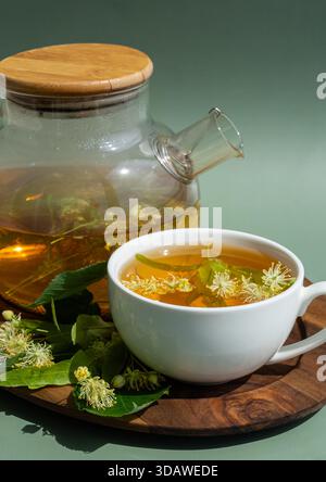Une théière en verre avec un couvercle en bois et une tasse blanche remplie de tisane tilleul. Des fleurs de tilleul frais sont dispersées sur une surface en bois, créant un cal Banque D'Images