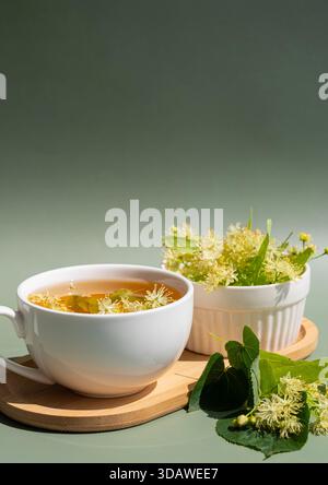 Une tasse en céramique blanche de thé tilleul à base de plantes avec des fleurs de tilleul frais et des feuilles vertes sur un plateau en bois. Le fond vert rehausse le thème naturel. Banque D'Images
