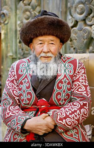 Kashgar, Chine - 06 décembre 2025 : vue d'un aîné avec un chapeau de fourrure et un manteau coloré à motifs, sa barbe aussi blanche que la neige d'hiver, un portrait de Banque D'Images