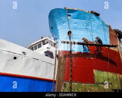 Dhaka, Bangladesh - 31 août 2024 : vue de travailleurs précaires perchés sur un échafaudage, peignant la coque d'un navire avec un bleu et un rouge vifs contre un cl Banque D'Images