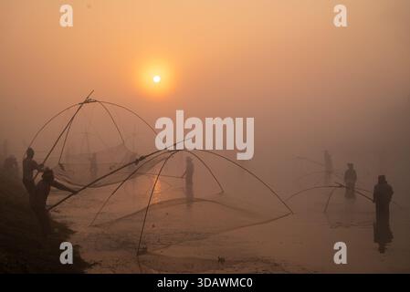 Pabna, Bangladesh - 29 novembre 2025 : vue sur la berge sereine où les silhouettes jettent des filets de pêche complexes dans l'aube dorée et brumeuse. Banque D'Images