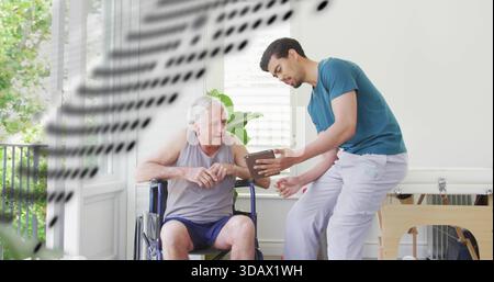 Tenant la tablette, soignant en T-shirt sarcelle guidant les aînés en fauteuil roulant à travers les exercices à la maison Banque D'Images