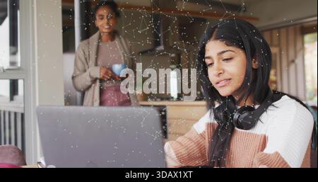 Taper fille adolescente dans le casque de chandail à l'aide d'un ordinateur portable à la table de cuisine mère tenant la tasse bleue regarder Banque D'Images