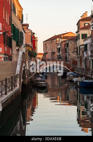 Lumière douce du soir sur le paisible canal du quartier Castello à Venise avec des maisons aux couleurs pastel, un pont en arc de brique et de petits bateaux reflétés dans l'eau calme. Banque D'Images