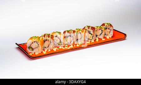 Rouleaux de sushi au saumon remplis de bâtonnets de crabe, d'avocat et de riz, garnis de sauce crémeuse et d'herbes fraîches, servis sur une assiette en céramique orange isolée. Banque D'Images