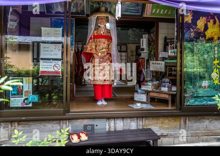Comme une mannequin de vitrine, Angela Berg se tient dans un kimono de style période Heian à la vitrine d'une boutique de location sur la route de pèlerinage Kumano Kodo à Nachikatsuura, préfecture de Wakayama, Japon. Des sandales en bois reposent sur un banc à l'extérieur, attendant le prochain client. Banque D'Images