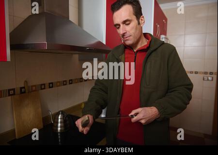 Un homme dans une chemise rouge et une veste verte prépare la nourriture dans une cuisine contemporaine, coupant avec un couteau à côté d'une bouilloire et d'une planche à découper, transportant à la maison Banque D'Images