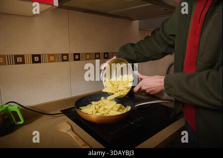 Une personne dans une veste verte verse des pommes de terre tranchées dans une poêle chaude sur une cuisinière, avec une cuillère en bois à proximité, représentant une cuisine maison chaude Banque D'Images