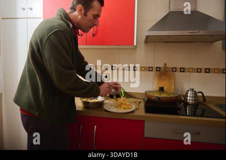 Un homme en veste verte prépare un repas dans une cuisine contemporaine. Il saupoudre du fromage sur des pâtes sur une assiette tandis qu'une casserole mijote sur le poêle. Banque D'Images