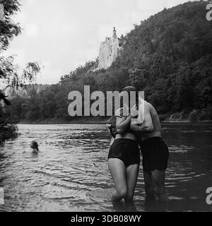 Slavyanogorsk (Svyatogorsk), Ukraine, URSS, années 1950 Une photo d'archives romantique en noir et blanc capturant un moment tendre sur la rivière Seversky Donets. Un jeune couple embrasse fermement tout en se tenant dans l'eau, profitant d'une journée d'été. En arrière-plan, l'église historique Saint-Nicolas se dresse au sommet des majestueuses falaises de craie. Cette image atmosphérique illustre de manière éclatante l'amour, les relations et les loisirs insouciants dans la paisible région du Donbass pendant l'ère soviétique. Banque D'Images