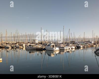 De nombreux voiliers et bateaux à moteur accostaient à une marina tranquille, leurs mâts se reflétant clairement dans l'eau calme sous un ciel lumineux Banque D'Images