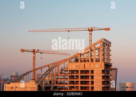 Structure à plusieurs étages inachevée baignée par la lumière chaude du coucher du soleil, avec des grues imposantes et un chantier de construction actif qui véhicule la croissance urbaine, eng Banque D'Images