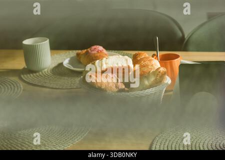 Un petit déjeuner chaleureux et accueillant avec un panier en osier de croissants et de pâtisseries, une tasse blanche et une tasse orange sur une table en bois, suggérant ho Banque D'Images