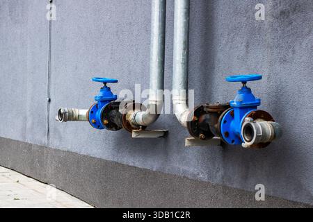 deux vannes d'eau bleues sur le mur montrant des tuyaux et des raccords en métal rouillé le long de l'extérieur en béton, supports boulonnés, écaillage Banque D'Images