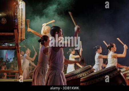Un groupe de batteurs de U-Theatre en robes traditionnelles blanches interprètent un ensemble de percussions rythmiques avec de grandes tambours Taiko sous les lumières de la scène. Banque D'Images