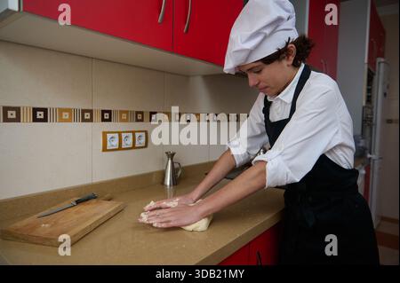 Un chef concentré en chemise blanche et tablier noir pétrit la pâte sur un comptoir beige, avec une planche à découper et un couteau à proximité dans une cuisine rouge vif. Banque D'Images