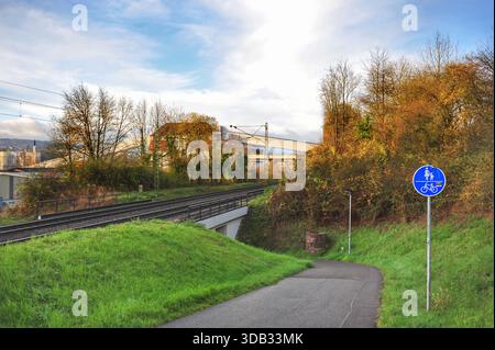 Piste cyclable à côté de Railway Under Clouds Blue Bicycle Sign indiquant Safe route le long de Wind Asphalt Grassy Embankment et Overhead Power Lines distantes Banque D'Images