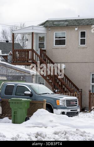 Canada, Québec, 29 novembre 2025 : poubelle verte, sol enneigé, camion GMC stationné, escalier arrière Banque D'Images