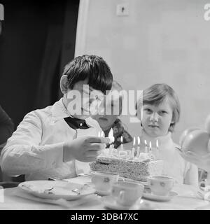 Sloviansk, Ukraine, URSS. 5 février 1977. Une photographie d'archives en noir et blanc en gros plan montre un garçon d'anniversaire dans une chemise blanche et un noeud papillon interagissant avec des bougies allumées sur un gâteau fait maison. Il se concentre intensément sur les flammes tandis que deux filles regardent avec curiosité. La table est sertie de tasses à thé et d'assiettes en porcelaine, capturant un moment franc d'une célébration d'enfance soviétique. L'image reflète la chaleur, l'innocence et les joies simples d'un rassemblement de famille dans la paisible région du Donbass au cours des années 1970 Banque D'Images