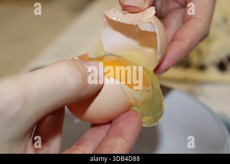 Une personne tient une coquille d'œuf dans une main tout en la cassant pour libérer le jaune et le blanc dans un bol. Cela a lieu dans un cadre de cuisine. Banque D'Images