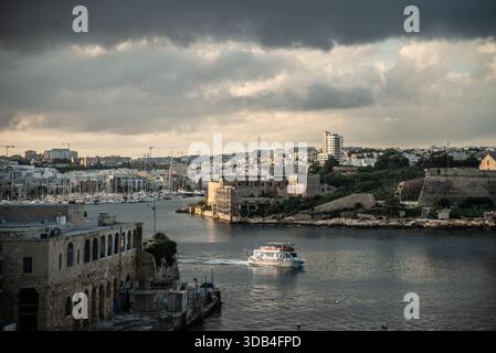 Un bateau passe par les criques du port de la Valette alors que le coucher de soleil se brise à travers les nuages sombres Banque D'Images