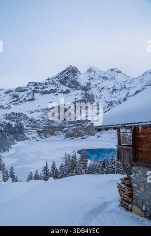 Cabane alpine enneigée surplombant un lac Oeschinen turquoise partiellement gelé dans les Alpes suisses Banque D'Images
