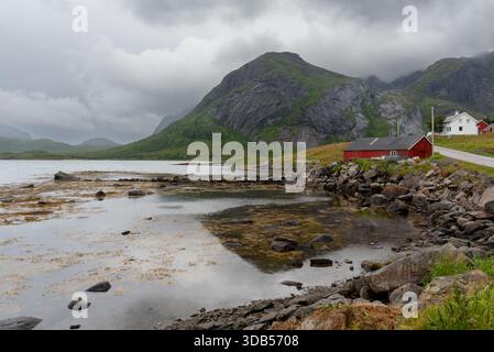 Rorbu rouge traditionnel debout sur la rive rocheuse d'un fjord lofoten sous un ciel couvert Banque D'Images