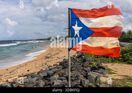 Drapeau de Porto Rico agitant sur la plage avec l'océan et le paysage côtier en arrière-plan. Banque D'Images