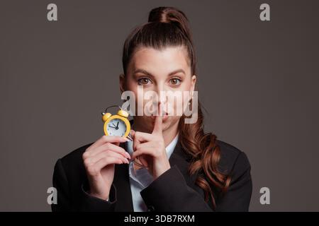 Femme en tenue d'affaires tenant un réveil jaune, faisant un geste de hush, mettant l'accent sur la gestion du temps, le silence et l'urgence. Banque D'Images